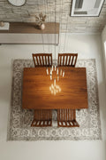 Wooden dining table with four chairs in a room with a patterned rug and stone wall.