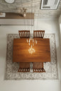 Wooden dining table with four chairs in a room with a patterned rug and stone wall.